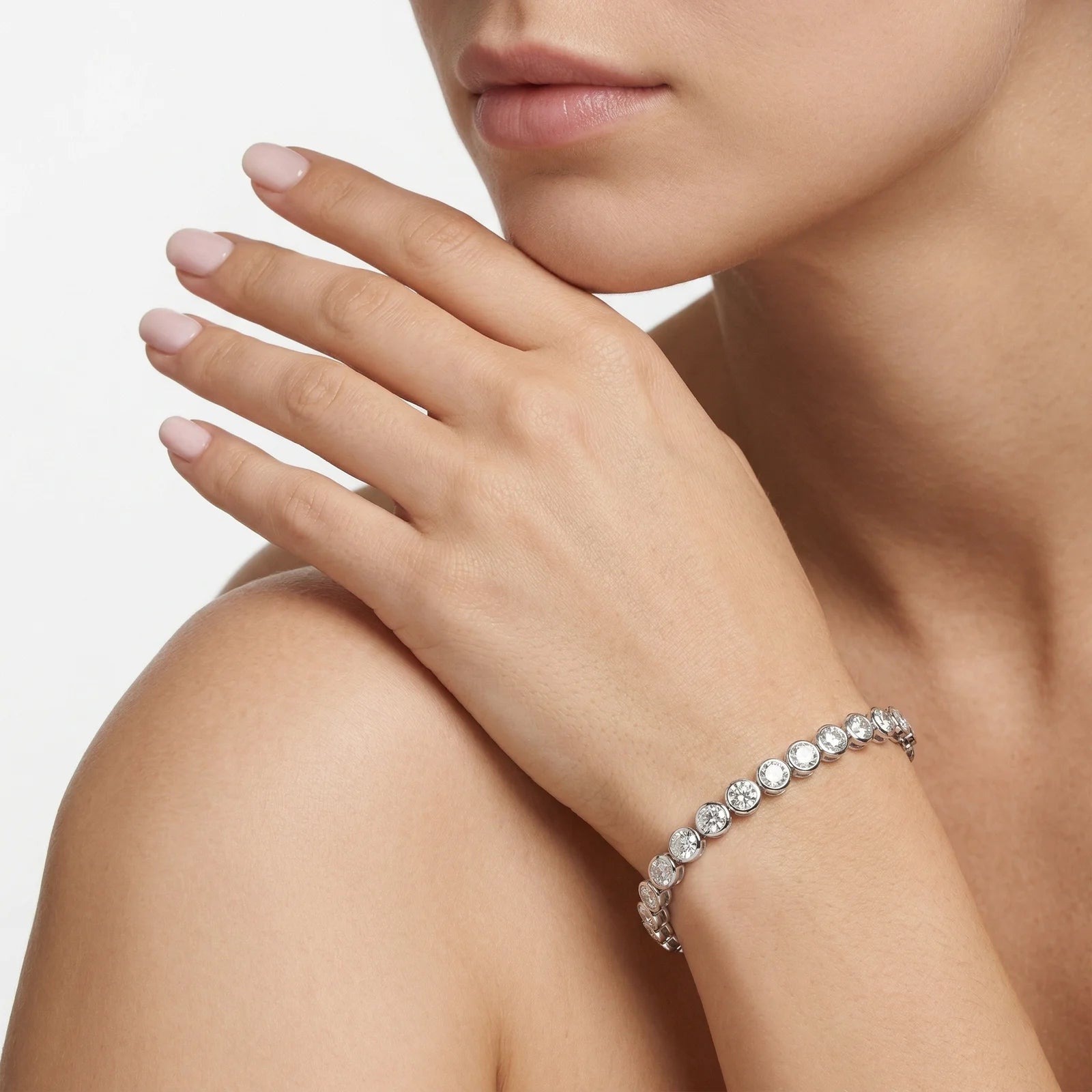 A close-up lifestyle shot of a woman wearing a silver-toned tennis bracelet with sparkling clear round-cut gemstones. The jewelry is shown on her wrist, with her hand resting near her chin against a clean, light gray background.
