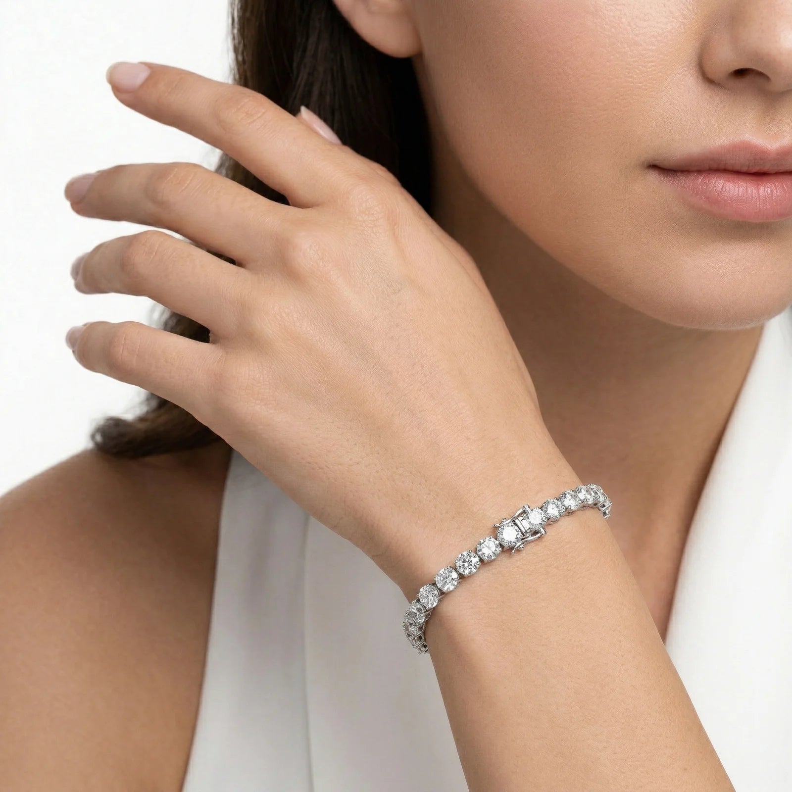 A close-up lifestyle shot of a model wearing a sparkling diamond tennis bracelet on her wrist, with her hand resting near her face against a clean white background.